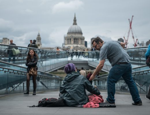 A homeless woman sits on the ground and a man hands her money. St Pauls Cathedral is in the background.